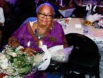 mary harris sits at table holding large bouquet and award