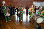 four african drummers in colorful african dress dance around large room